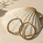 Grote gouden twisted hoops Chloe Earrings van hi SISU tegen een schelp op het strand
