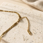 Close-up van de sluiting en het snake-patroon van de goudkleurige Taylor Necklace van hi SISU, flatlay op travertin
